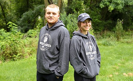 Two smiling people, wearing grey hoodies, stand back to back with their arms crossed.